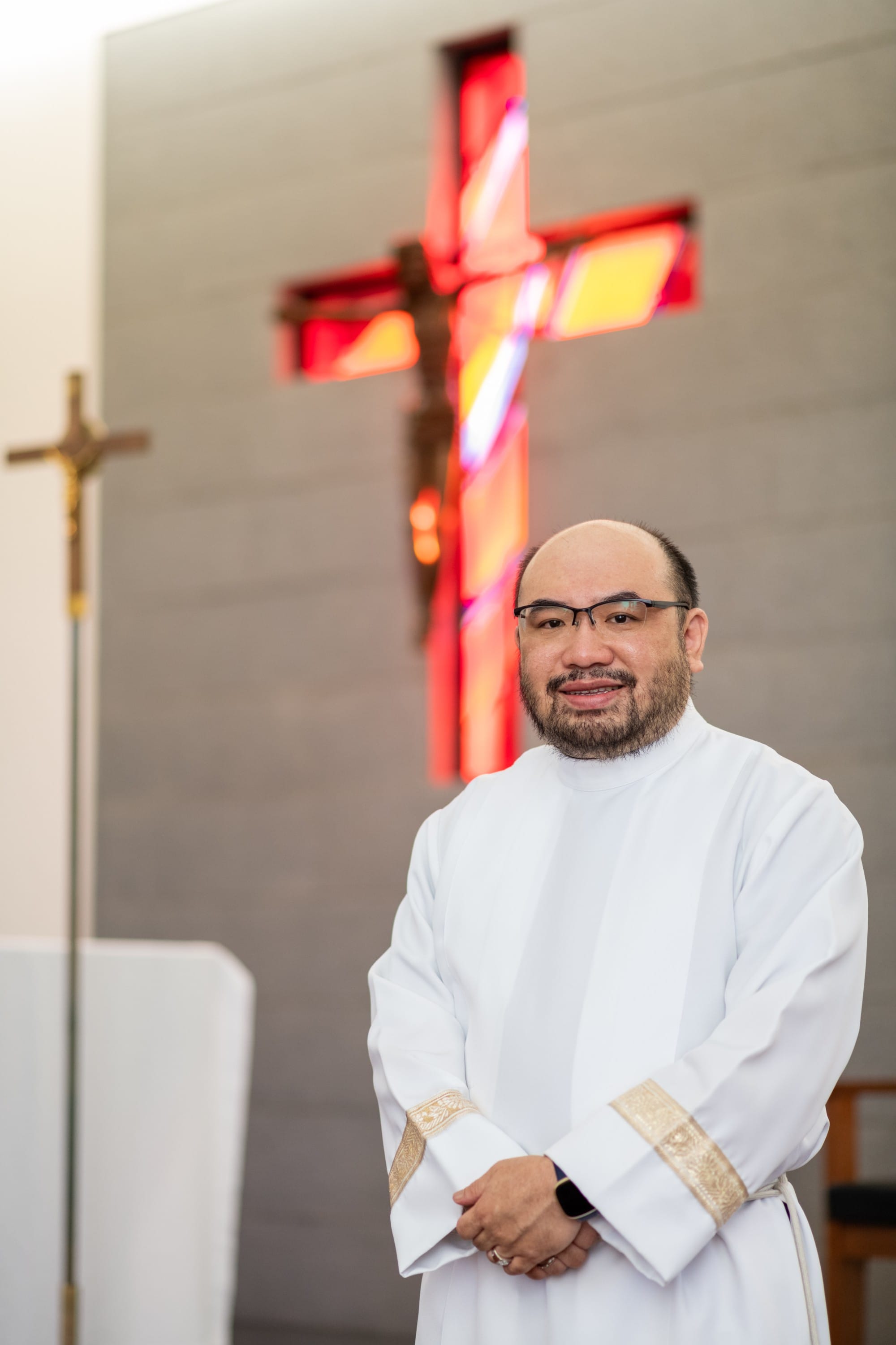 A smiling man in white clergy robes stands before a large illuminated red stained glass crucifix on a church wall.